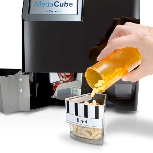 A hand pouring a large amount of pills into a large clear MedaCube bin. The restock door is open on the MedaCube showing the inside of the device.