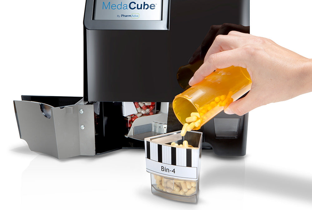 A hand pouring a large amount of pills into a large clear MedaCube bin. The restock door is open on the MedaCube showing the inside of the device. 