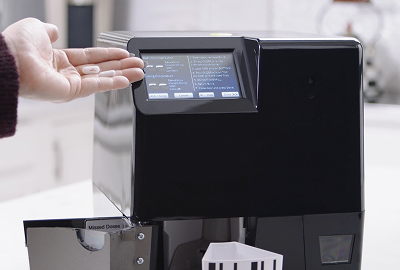 MedaCube on a counter showing one of the refill screens with the restock door open and bin pulled out. A hand is holding a pill that matches the medication on screen.