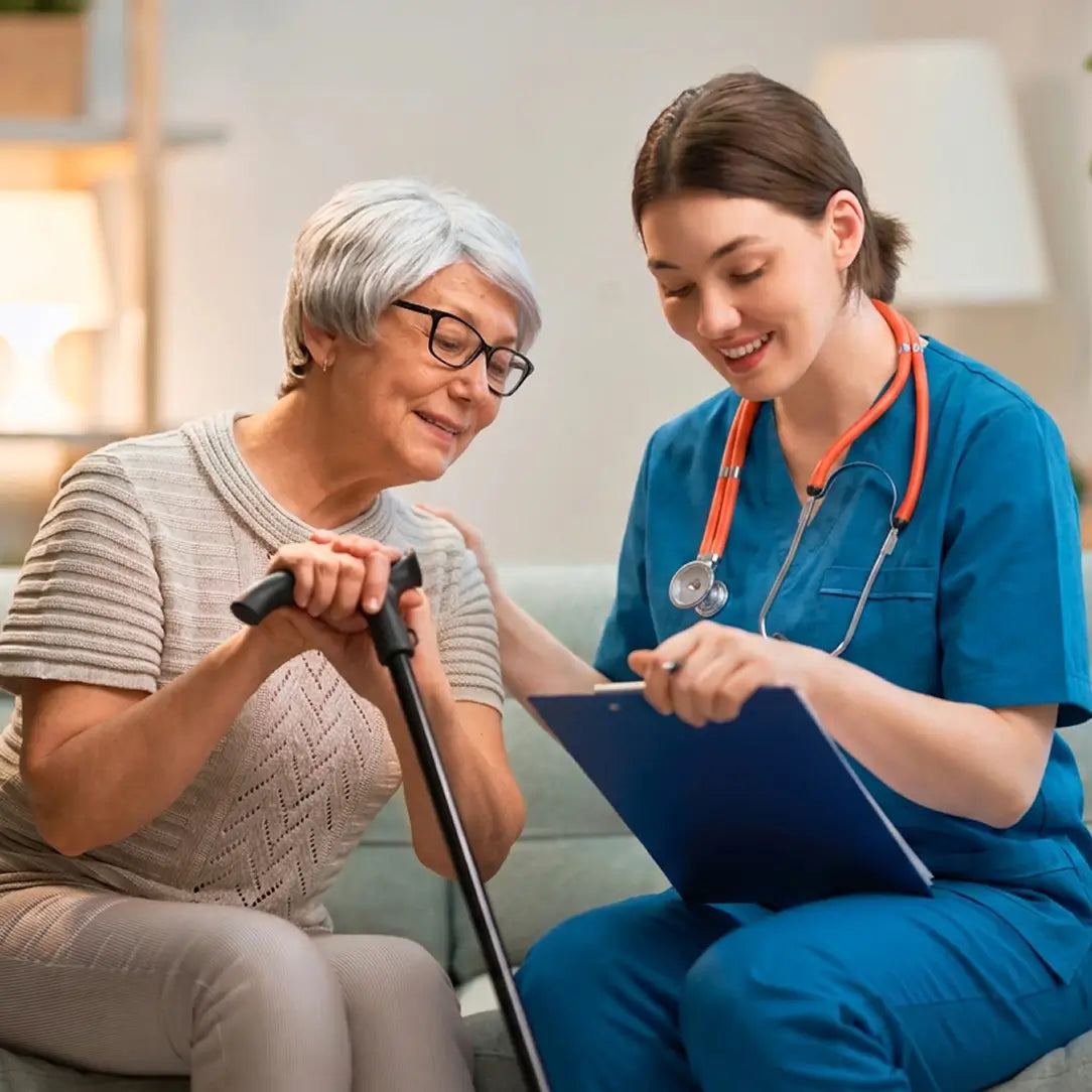 A young female healthcare worker in blue scrubs with an orange stethoscope smiles while holding a clipboard next to an elderly woman with gray hair and glasses who is using a cane in a brightly lit indoor setting.
