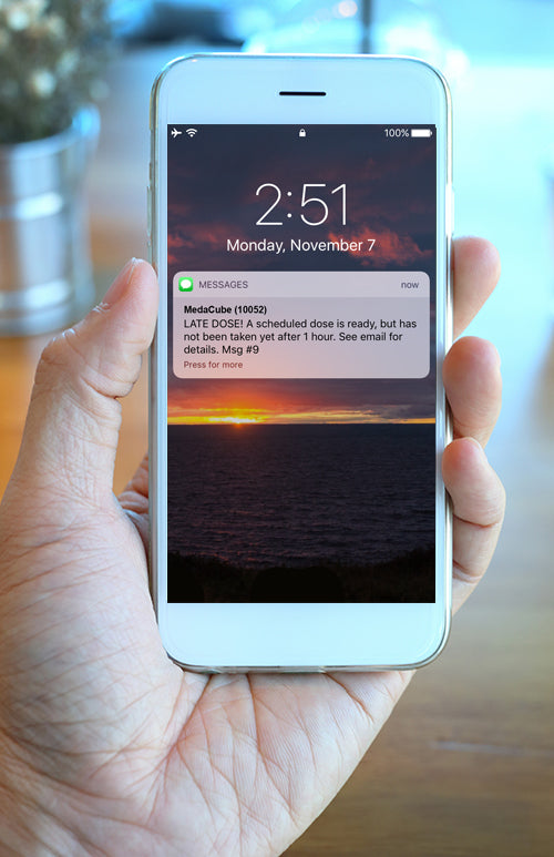A hand holding a phone. On the phone there is a text notification regarding a late dose from the MedaCube.